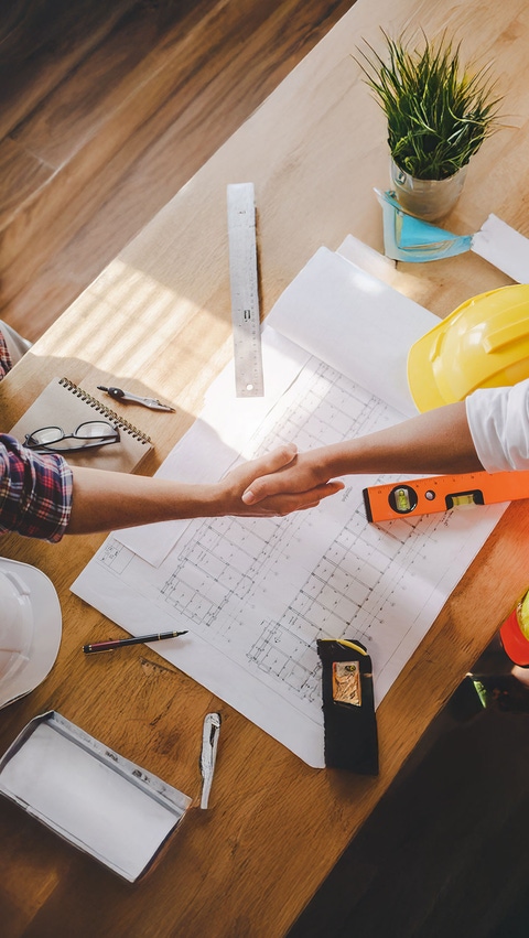 Deux personnes se serrant la main au-dessus d’une table exposant des dessins et des outils de construction. Deux personnes se serrant la main au-dessus d’une table exposant des dessins et des outils de construction.