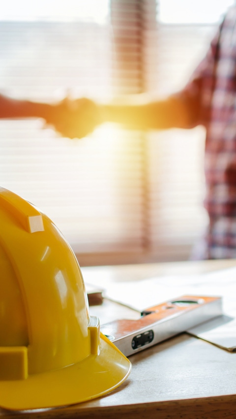Image of a table with construction plays and hard hat with two people shaking hands in the background Image of a table with construction plays and hard hat with two people shaking hands in the background